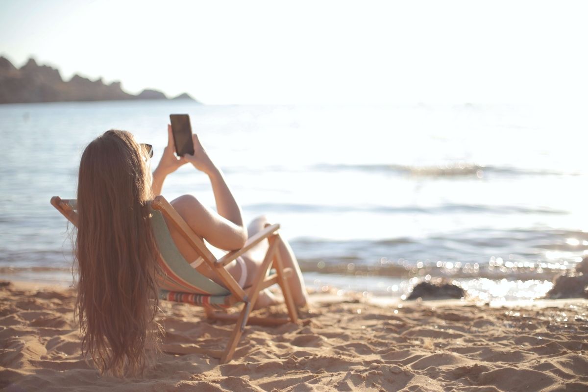 mulher a mexer no telemóvel sentada em cadeira na praia a apanhar sol
